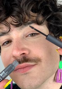 Mascara à sourcils dans un emballage gris étiqueté "972" tenu dans une main avec une brosse applicateur noire ; le modèle a des cheveux bouclés et une moustache.