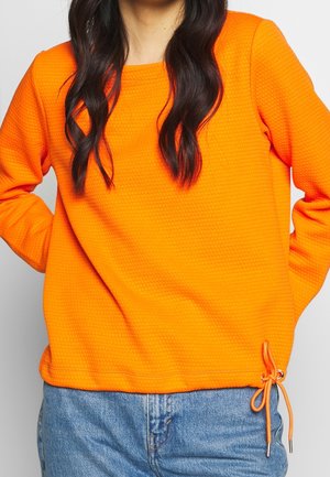 Kobieta w jasnopomarańczowej, teksturowanej bluzce z długim rękawem i sznurkiem regulującym na dole, w połączeniu z jasnoniebieskimi dżinsami.