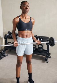 Juoda sportinė liemenėlė su logotipu, šviesiai mėlyni treniruoklių šortai, laikanti štangą. Sporto salė su hanteliais ir kettlebell'ais fone.