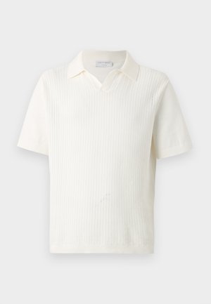 Bărbat tânăr cu păr creț purtând un polo tricotat alb cu mânecă scurtă, pantaloni scurți kaki, șosete albe și adidași albi, stând pe un fundal simplu.