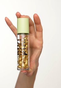 Bouteille en verre transparent avec un bouchon vert, remplie de capsules dorées. Le texte noir à l'avant indique "Pixi SkinTreats."