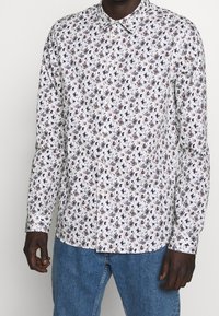 Bílá košile s krátkým rukávem a květinovým vzorem v tmavě modré a burgundské barvě, dlouhé rukávy a klasický límec. Nosená s modrými denimovými kalhotami.