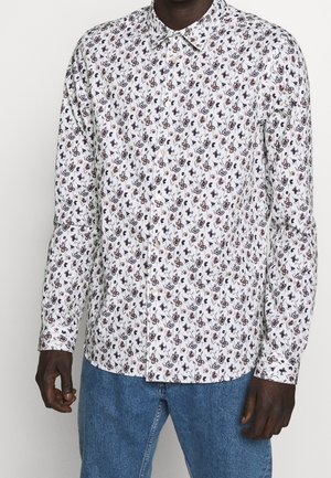 Camicia bianca con motivo floreale in blu scuro e bordeaux, maniche lunghe e colletto classico. Indossata con pantaloni di denim blu.