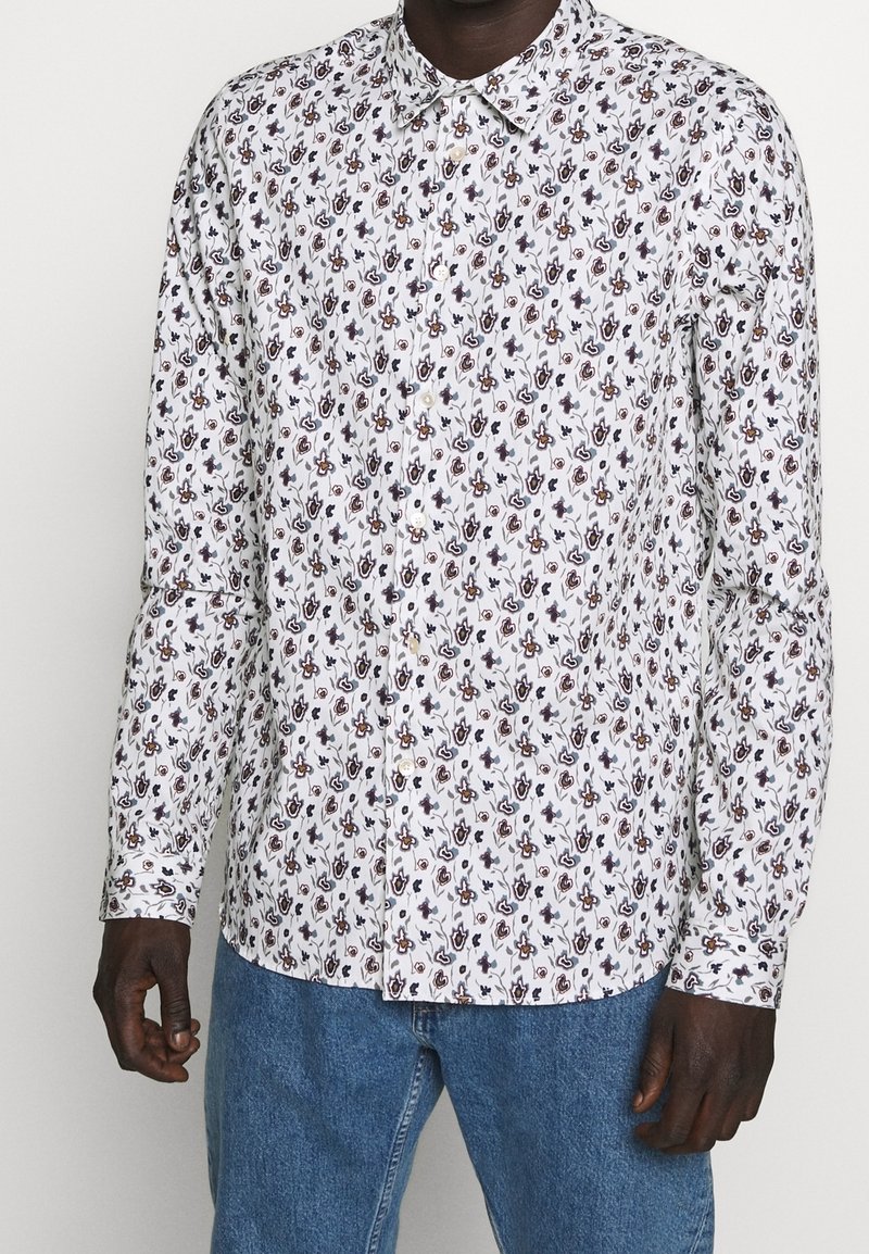 Bílá košile s krátkým rukávem a květinovým vzorem v tmavě modré a burgundské barvě, dlouhé rukávy a klasický límec. Nosená s modrými denimovými kalhotami.