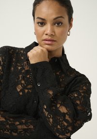 Zwarte kant blouse met een kraaghalslijn, knoopsluiting en lange mouwen met ingewikkelde bloemmotieven en een doorschijnende textuur.