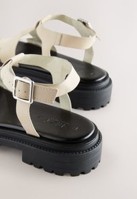 Sandale cu bretele bej și talpă platformă groasă de culoare neagră. Prezintă o baretă reglabilă cu cataramă și bretele moi pentru confort.
