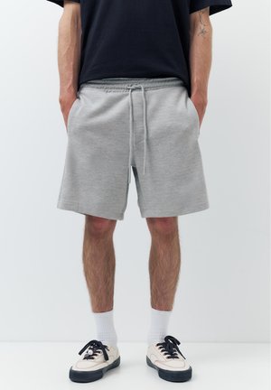 Homme portant un short gris à cordon, des chaussettes blanches montantes et des baskets blanches avec semelles et lacets noirs, debout les mains dans les poches.