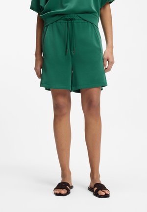 Henkilö, jolla on vihreät kiristettävät shortsit ja vastaava yläosa, yhdistettynä tummanruskeisiin avonaisiin liukusandaaleihin, seisoo valkoisen taustan edessä.