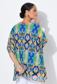 Blusa trasparente a fantasia colorata con tonalità di blu, verde, giallo e arancione; vestibilità ampia con maniche larghe e orlo asimmetrico.
