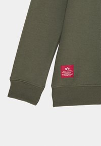 Felpa a maniche lunghe verde militare con polsini e orlo a coste, caratterizzata da un piccolo patch rettangolare rosso con il logo Alpha Industries vicino all'orlo inferiore.