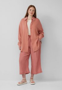 Chemise boutonnée rose poussiéreuse avec une texture côtelée, assortie à un pantalon court ample et loose, et des sandales blanches à lanières, sur un fond neutre.
