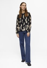 Blusa de estampado negro y crema con mangas largas y cuello redondo, combinada con jeans acampanados de cintura alta en azul y zapatos negros.