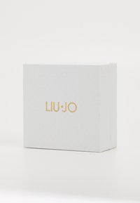 Balts teksturēts kaste ar zelta "LIU·JO" logo centrā priekšpusē uz vienkārša balta fona.