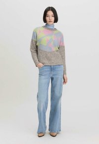 Gehaakte trui in grijs met multiculturele geometrische patronen (roze, groen, blauw) gecombineerd met lichtblauwe flared jeans en bruine hakken.