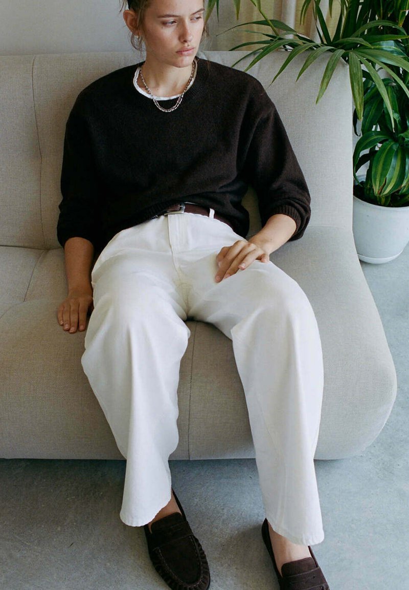 Jeune personne vêtue d'un pull sombre et d'un pantalon blanc assise sur un canapé clair à côté d'une grande plante verte dans un pot blanc.
