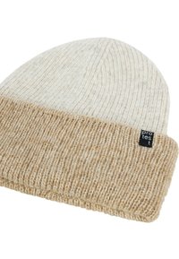 Two-tone knit beanie with beige folded cuff and light cream top, featuring a small black rectangular brand tag on the cuff.