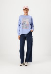 Lichtblauw sweatshirt met grafische print, ruime pasvorm, gecombineerd met donkerblauwe wijde spijkerbroek en grijze sneakers, afgewerkt met een roze pet.