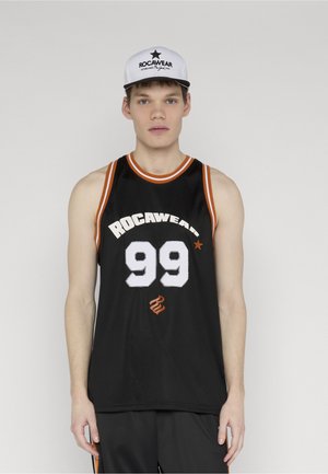 Noor mees mustas Rocawear korvpallivestiga, millel on number 99, ning valge Rocawear logoga nokamütsuga ühevärvilise tausta ees.