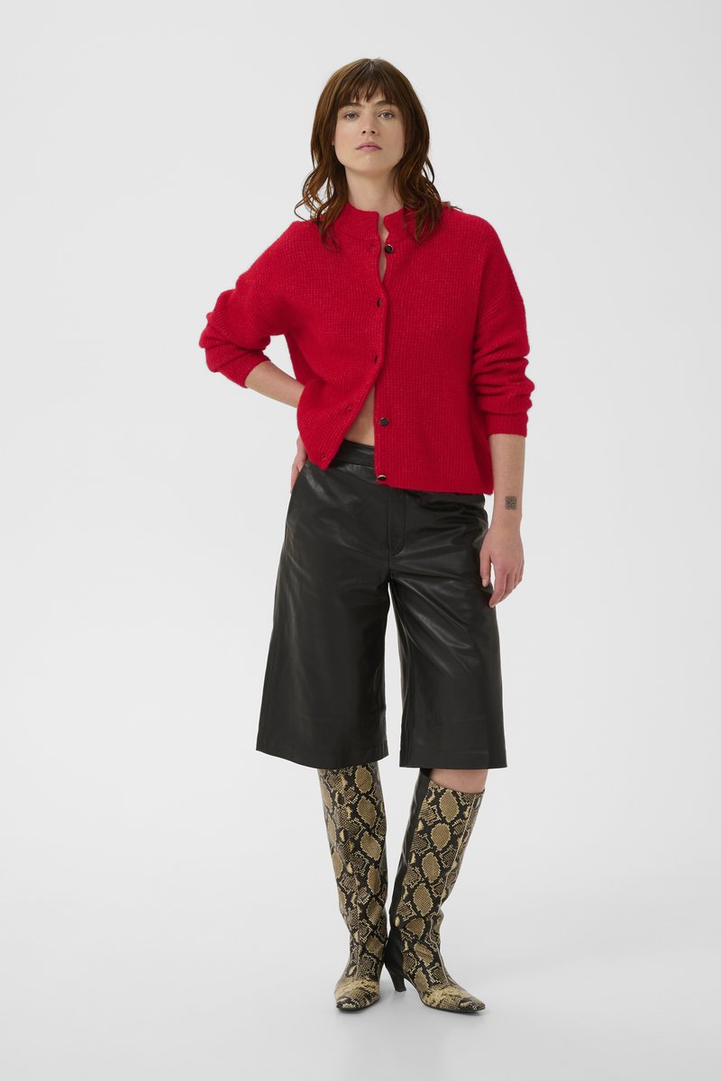 Femme portant un cardigan rouge boutonné, un short en cuir noir longueur genou et des bottes montantes avec un motif peau de serpent, debout, une main sur la hanche.