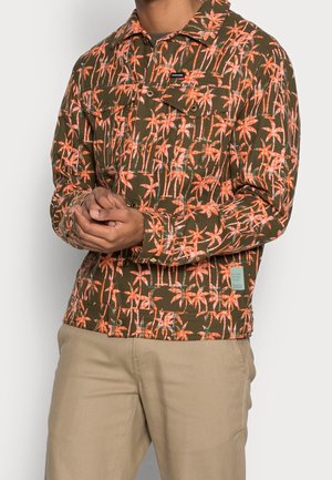 Man wearing olive green jacket with bright orange palm tree pattern and beige pants, hands clasped in front.