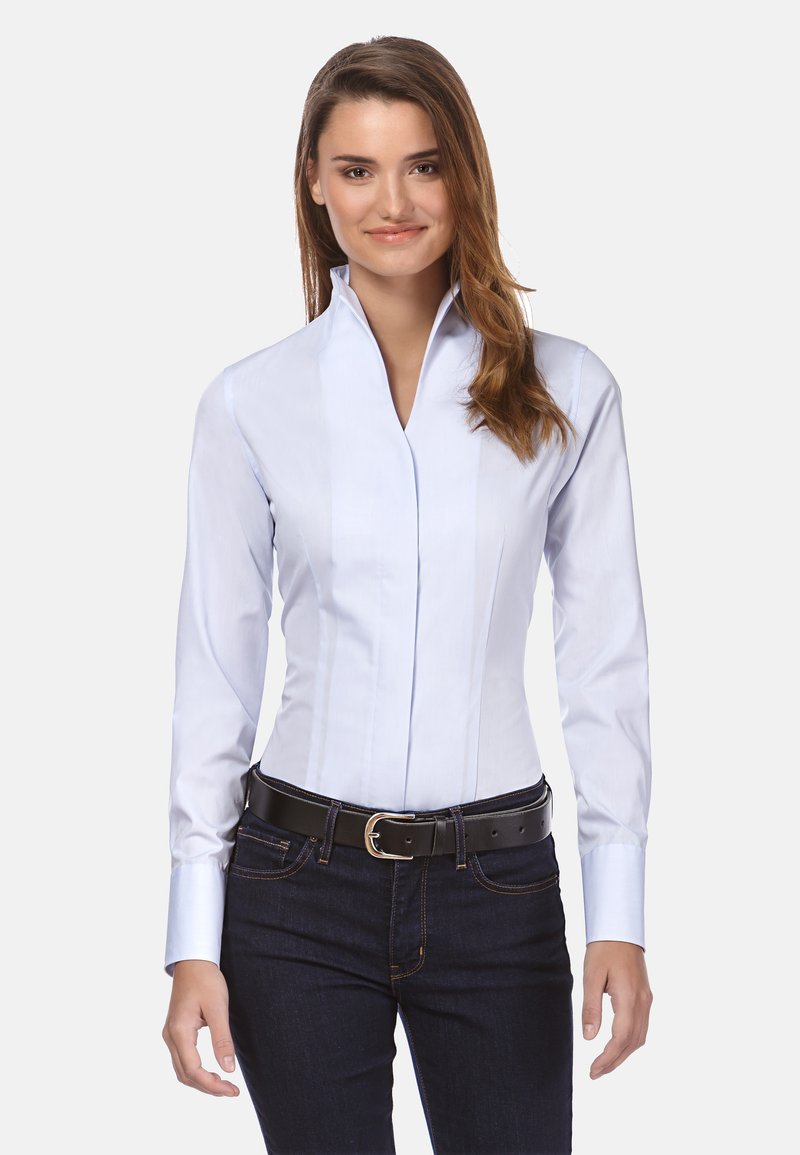 Chemise habillée à manches longues bleu clair avec un col haut, présentant des coutures verticales et une texture lisse, assortie à un jean sombre et une ceinture noire.