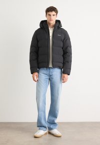 Zwarte gewatteerde jas met een capuchon, voorzien van een rits aan de voorkant en een logo op de borst. Draagt in combinatie met lichtblauwe jeans en witte sneakers.