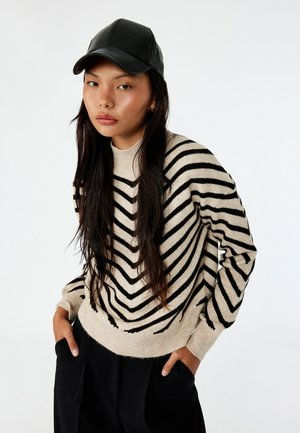 Maglione beige con strisce a zigzag nere, colletto rotondo e maniche lunghe. Indossato con pantaloni neri e un cappello di pelle nera.