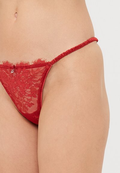 Red lace thong with thin elastic sides, floral pattern along the front, adorned with a small decorative detail at the center.