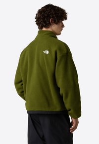 Chaqueta de forro polar verde oliva con cuello alto, con un dobladillo negro en la parte inferior y un logo blanco en la parte trasera. Apariencia texturizada y cálida.