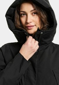 Schwarze isolierte Jacke mit hohem Kragen und Kapuze, ausgestattet mit einer glatten Textur und Druckknöpfen. Nahaufnahme einer Hand auf der Jacke.