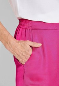 Fitted rozen satijnen broek met een elastische tailleband, voorzien van een zijzak en een zachte textuur. Close-up van een hand die op de zak rust.