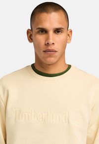 Sweatshirt crème en coton avec un logo texturé "Timberland" et un design d'arbre, doté d'un col côtelé vert en dessous.