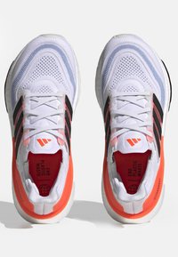 Białe buty sportowe z siateczkową cholewką, z czarnymi paskami, jaskrawymi koralowymi akcentami i szarą podeszwą. Tekst wewnątrz brzmi "ZAKOŃCZ ZANIECZYSZCZANIE PLASTIKIEM."