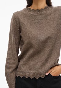 Smeđi pleteni pulover s teksturiranim, valovitim izrezom i helikopter uzorkom na rubu. Dugi rukavi i uski kroj.