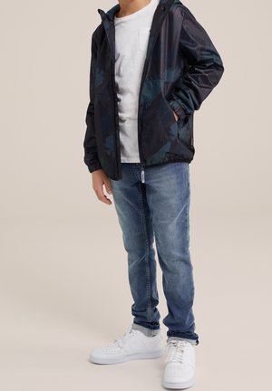 Windbreaker jacket in dark blue with a geometric pattern, paired with a white t-shirt, blue jeans, and white sneakers.
