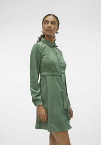 Robe en velours côtelé vert avec des manches longues, une fermeture à boutons à l'avant et une ceinture à la taille. Présente des rayures verticales et un col classique.