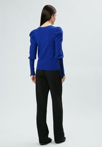 Blauer Strickpullover mit puffartigen Ärmeln und gerippten Bündchen, kombiniert mit schwarzen maßgeschneiderten Hosen. Auffälliger Textdetails am Ärmel lautet "INFLUENCER."
