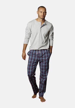 Man walking barefoot wearing a light gray long-sleeve henley shirt and blue and red plaid pajama pants against a plain background.