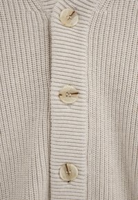 Nahaufnahme eines beigen, gestrickten Stoffs mit vertikaler Rippenstruktur und drei durchscheinenden, runden Knöpfen, die in einer vertikalen Linie angenäht sind.