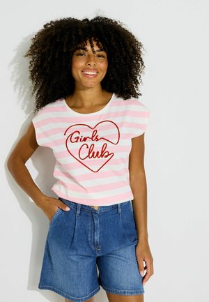 Mujer sonriente con cabello rizado que lleva una camiseta de rayas rosa y blanca con "Girls Club" dentro de un corazón y shorts vaqueros azules, está apoyada contra la pared.