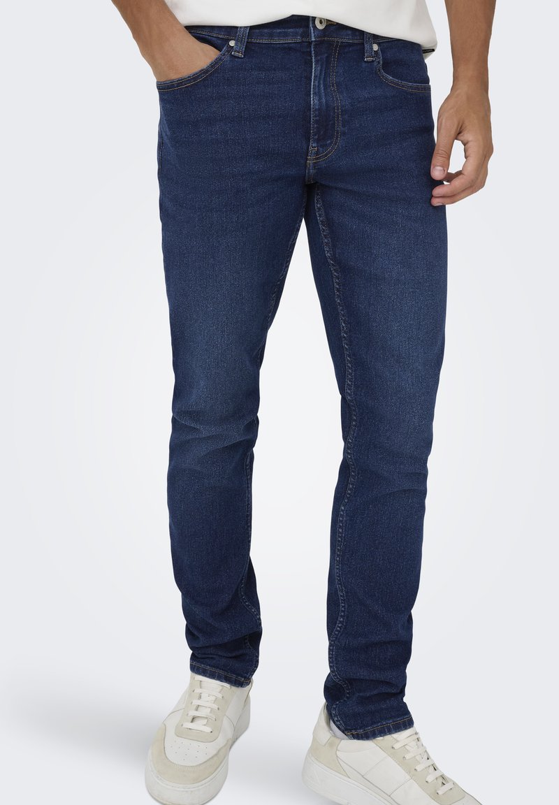 Jean en denim bleu à coupe slim, avec un design à cinq poches, des surpiqûres discrètes et une jambe droite se terminant au-dessus de baskets blanches.