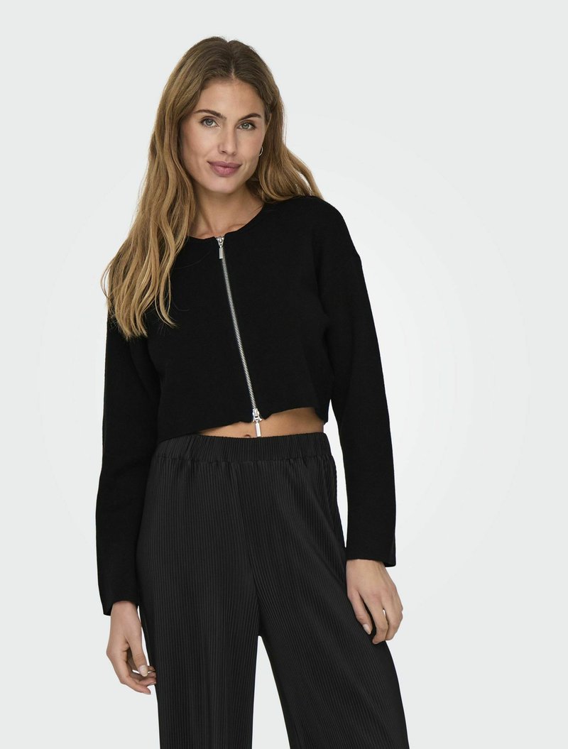 Maglione nero corto con zip frontale, abbinato a pantaloni larghi plissettati neri. Texture morbida, design minimal e vestibilità rilassata.
