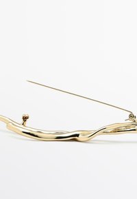 Fermaglio per capelli in metallo color oro, con una forma organicamente attorcigliata e una punta appuntita, dotato di una chiusura per un fissaggio sicuro. Superficie riflettente.