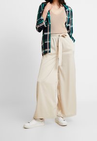 Béžové kalhoty s širokými nohavicemi a zavázaným pasem, zkombinované s světle béžovým topem s výstřihem do V a otevřenou bundou v zeleno-námořnickém kostkovaném vzoru. Bílé tenisky doplňují tento outfit.