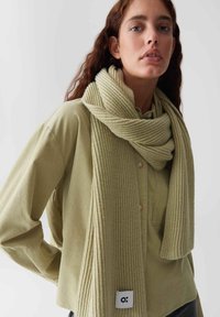 Gebreide sjaal in lichtgroen met ribbeltextuur, om de nek gedrapeerd; bevestigd label toont logo. Draagt een bijpassende groene shirt.