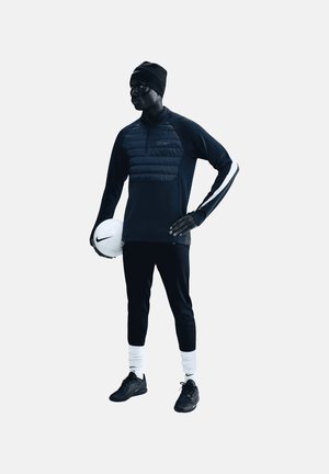 Homme en tenue de sport noire et casquette tient un ballon de football blanc Nike d'une main, l'autre main posée sur la hanche, debout devant un fond uni.