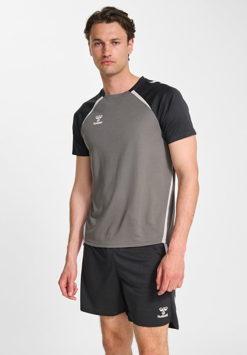 Mann steht und trägt ein graues und schwarzes kurzärmeliges Sportshirt sowie schwarze Shorts mit Hummel-Logos auf weißem Hintergrund.