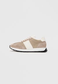 Beige semsket sko med en hvit lærstripe, snøring foran, polstret krage og svart gummisåle. Minimalistisk design, lett konstruksjon.