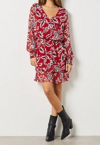 Vestido rojo con estampado floral, mangas largas y transparentes, escote en V, cintura ajustada y dobladillo con volantes. Llevado con botas negras de tobillo.
