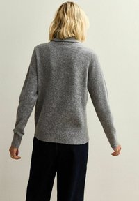 Grå ulltröja med hög krage, ribbade muddar och rak silhuett, matchad med mörka byxor mot en neutral bakgrund.